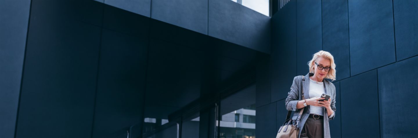 Una mujer de negocios rubia con blazer gris está frente a un edificio moderno, mirando su smartphone y sonriendo.