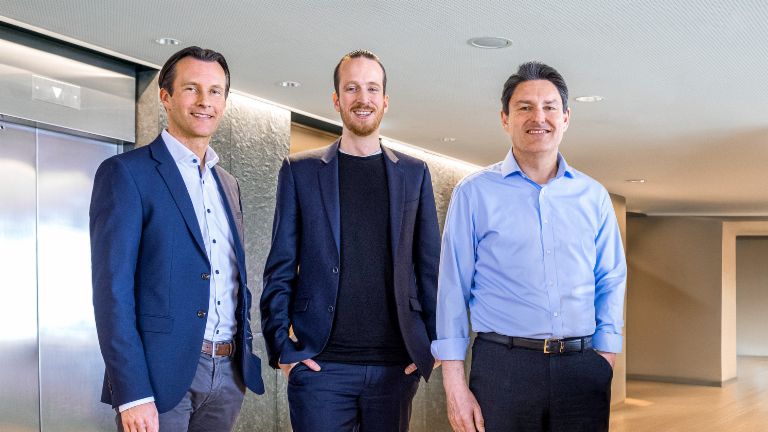Tres miembros masculinos de la Junta Directiva del Grupo EOS están de pie en traje de negocios frente a ascensores en un entorno moderno.