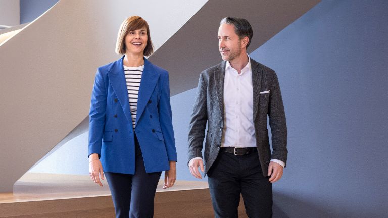 El CEO en un blazer gris y la CFO del Grupo EOS en un blazer azul caminan por una oficina moderna. El espacio cuenta con grandes ventanas, una pared curva y elegantes lámparas colgantes.