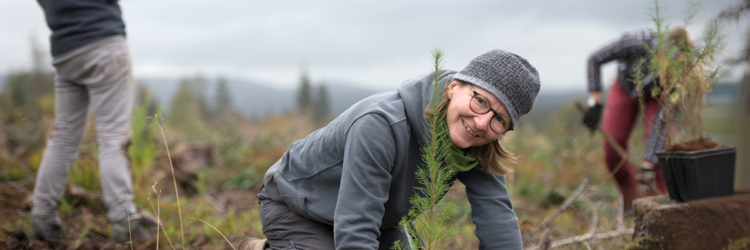 Una mujer joven con sudadera, pantalón y gorro grises está arrodillada en un campo delante de un árbol recién plantado y sonríe a la cámara. Al fondo, a la derecha, distinguimos a otra persona con pantalón rojo cavando un agujero para plantar un árbol. En el extremo izquierdo de la imagen, vemos la imagen cortada de alguien que sostiene un teléfono móvil y está a punto de hacer una foto.