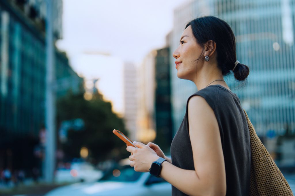 Una joven está de pie al aire libre en un entorno urbano, sostiene su smartphone y sonríe mirando hacia adelante. Al fondo se ven edificios modernos y árboles.