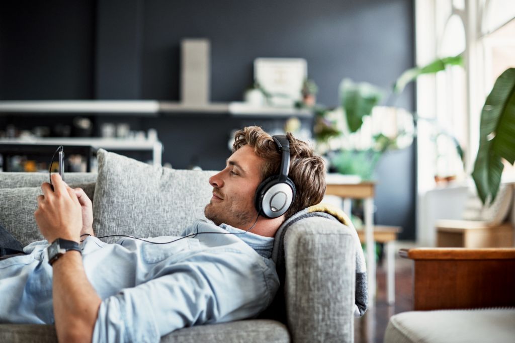 Un joven está tumbado en un sofá en su sala de estar, lleva auriculares y mira su smartphone sonriendo, disfrutando de música o un podcast.