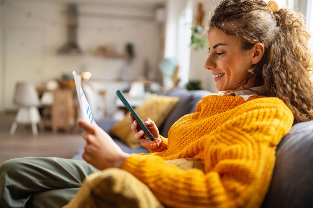 Una joven sonriente con el pelo rizado, vestida con un acogedor jersey amarillo, se sienta en un sofá en su sala de estar y mira su smartphone mientras sostiene un documento de papel.