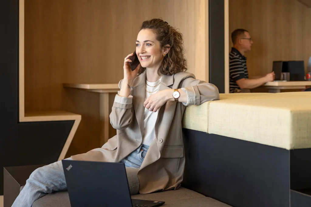 Una persona sonriente está sentada en una moderna zona lounge de oficina, hablando por teléfono y trabajando en un portátil. Al fondo, otras dos personas están trabajando.