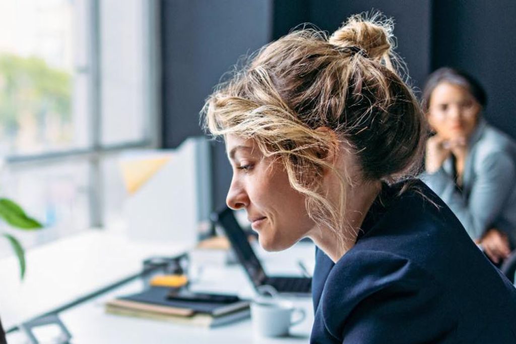 La imagen muestra a una mujer sentada en un escritorio en una oficina moderna, vistiendo un blazer oscuro mientras se concentra en su laptop, con una planta y algunos suministros de oficina visibles en la mesa, y otra persona sentada en el fondo.