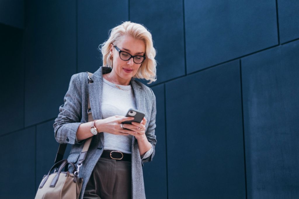 Una mujer de negocios rubia con blazer gris está frente a un edificio moderno, mirando su smartphone y sonriendo.