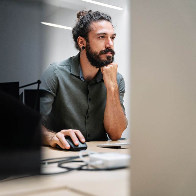 Un hombre con barba y el cabello largo recogido en un moño está sentado frente a un ordenador, completamente absorto en su trabajo.