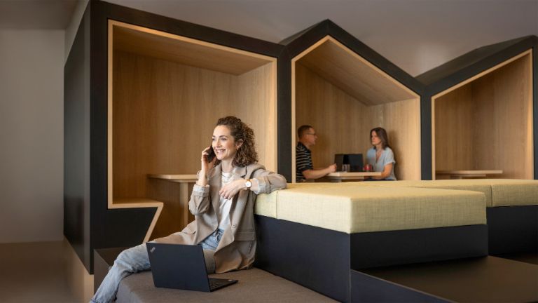 Una persona sonriente está sentada en una moderna zona lounge de oficina, hablando por teléfono y trabajando en un portátil. Al fondo, otras dos personas están trabajando.
