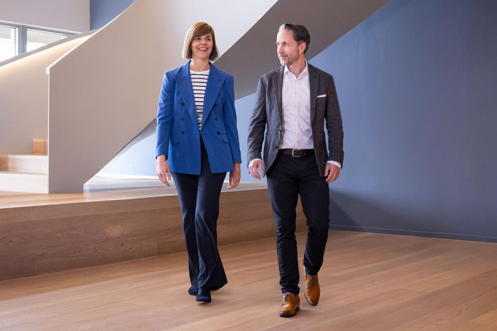 El CEO en un blazer gris y la CFO del Grupo EOS en un blazer azul caminan por una oficina moderna. El espacio cuenta con grandes ventanas, una pared curva y elegantes lámparas colgantes.