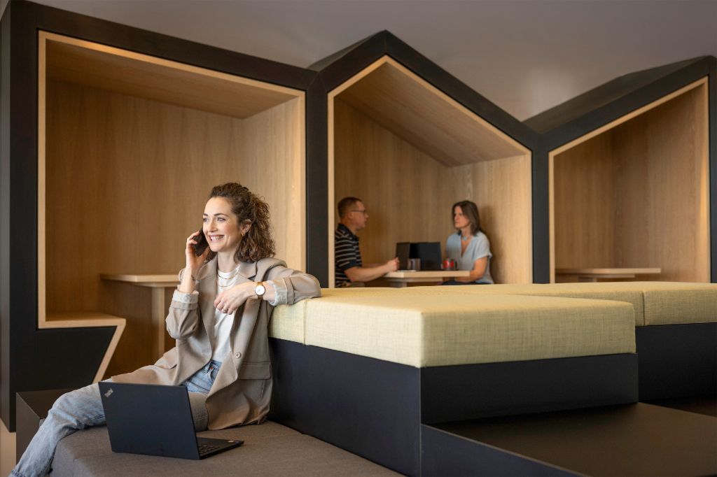 Una persona sonriente está sentada en una moderna zona lounge de oficina, hablando por teléfono y trabajando en un portátil. Al fondo, otras dos personas están trabajando.