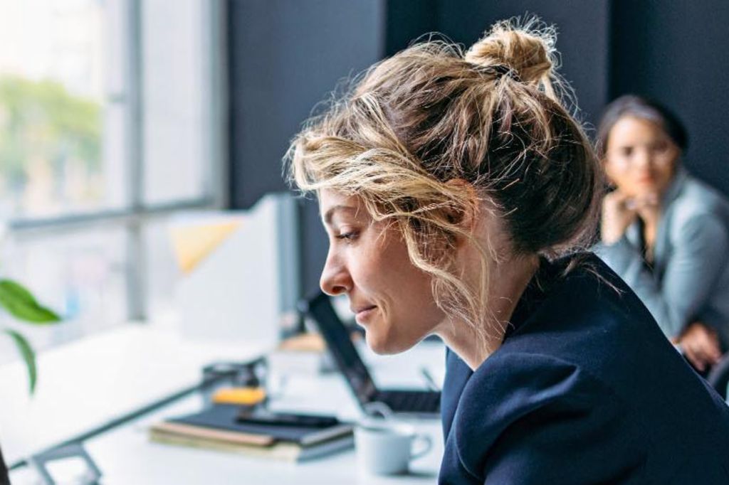 La imagen muestra a una mujer sentada en un escritorio en una oficina moderna, vistiendo un blazer oscuro mientras se concentra en su laptop, con una planta y algunos suministros de oficina visibles en la mesa, y otra persona sentada en el fondo.