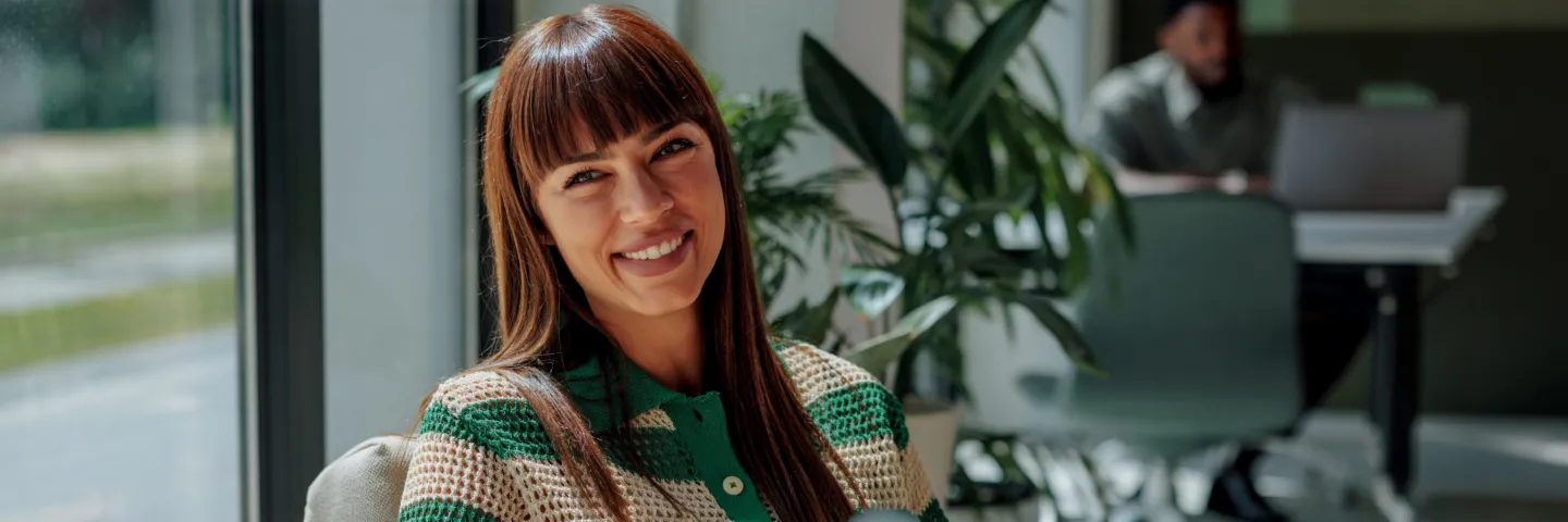 Una mujer sonriente de cabello oscuro está sentada cómodamente en una oficina moderna y luminosa y mira con calidez a la cámara. Al fondo se ven otras personas y grandes ventanales.