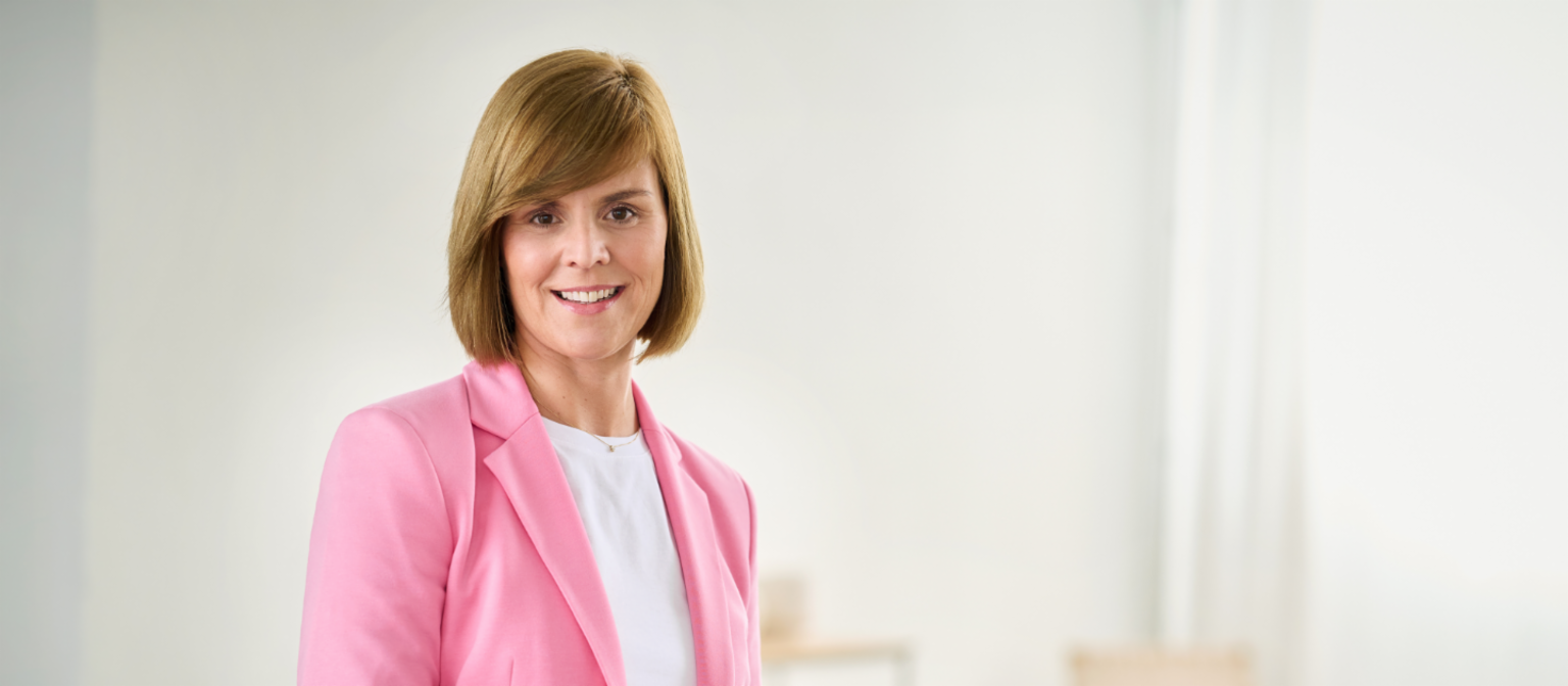 Eva Griewel, mujer vestida con americana de color rosa y camiseta blanca de pie en una estancia luminosa