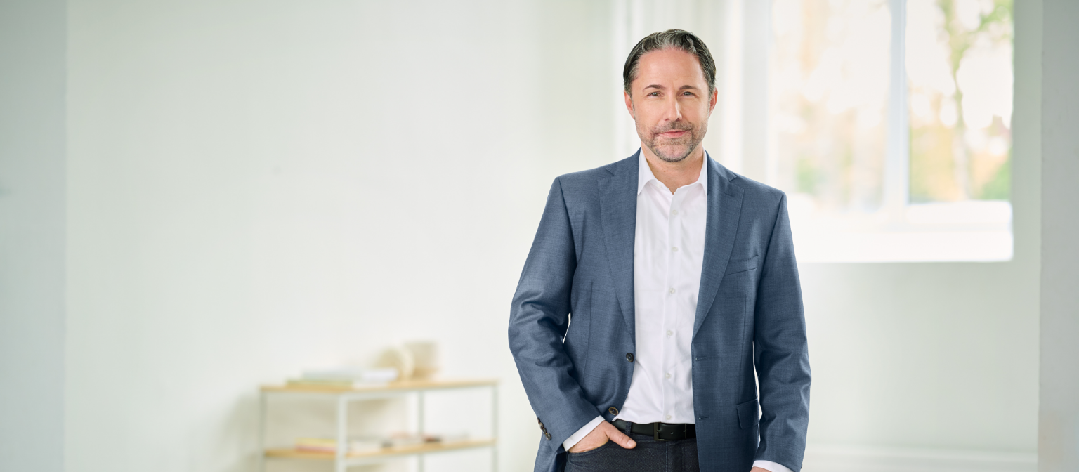 Marwin Ramcke, CEO se encuentra de pie vestido con un traje azul y la mano derecha en el bolsillo del pantalón en una estancia luminosa. 