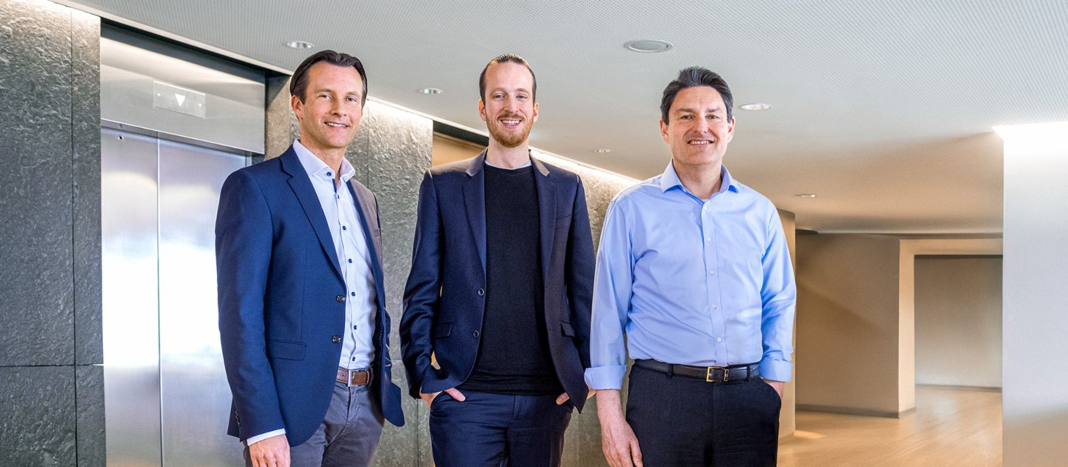 Tres miembros masculinos de la Junta Directiva del Grupo EOS están de pie en traje de negocios frente a ascensores en un entorno moderno.