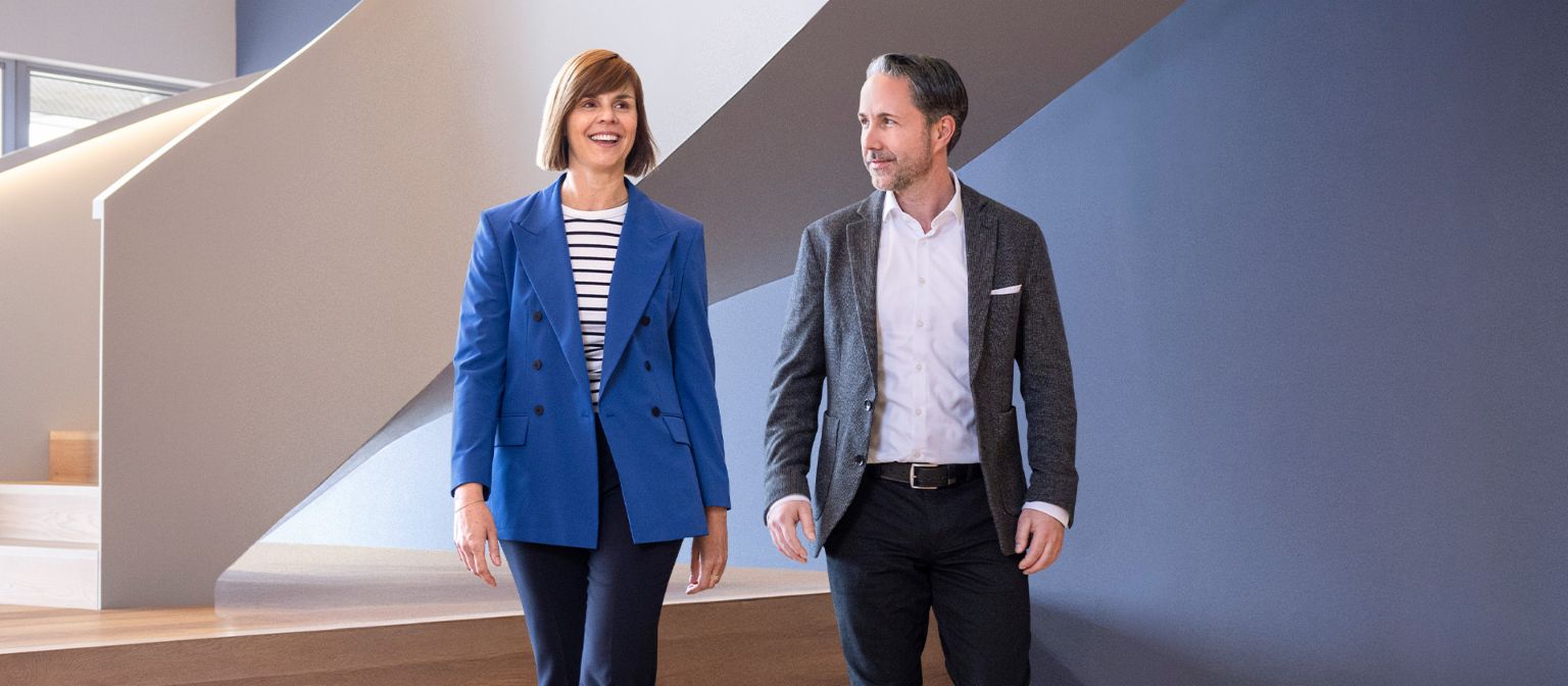 El CEO en un blazer gris y la CFO del Grupo EOS en un blazer azul caminan por una oficina moderna. El espacio cuenta con grandes ventanas, una pared curva y elegantes lámparas colgantes.