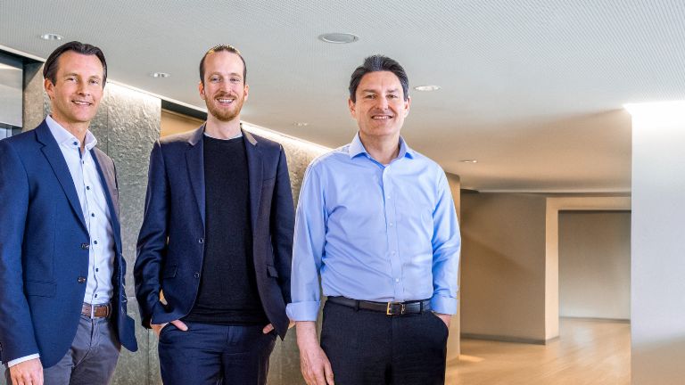 Tres miembros masculinos de la Junta Directiva del Grupo EOS están de pie en traje de negocios frente a ascensores en un entorno moderno.