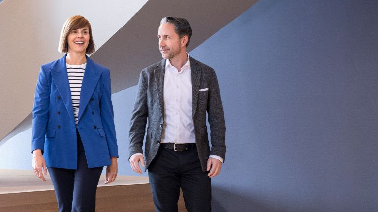 El CEO en un blazer gris y la CFO del Grupo EOS en un blazer azul caminan por una oficina moderna. El espacio cuenta con grandes ventanas, una pared curva y elegantes lámparas colgantes.