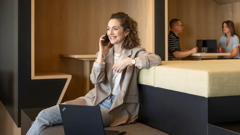 Una persona sonriente está sentada en una moderna zona lounge de oficina, hablando por teléfono y trabajando en un portátil. Al fondo, otras dos personas están trabajando.