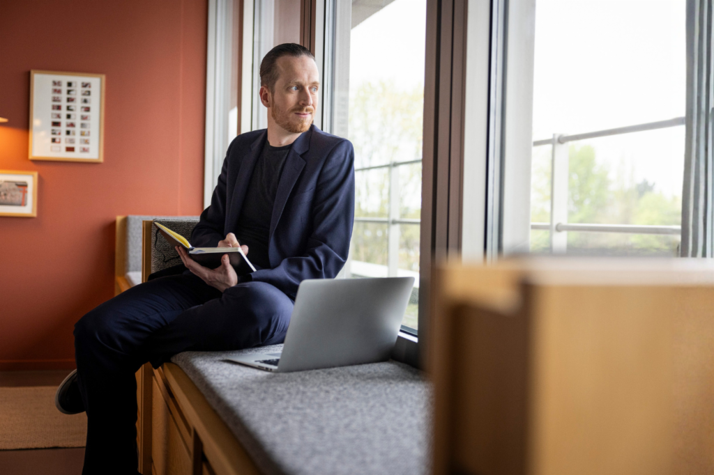 Sebastian Pollmer está sentado en el alféizar de una ventana con un portátil abierto y una libreta en la mano, mirando por la ventana.