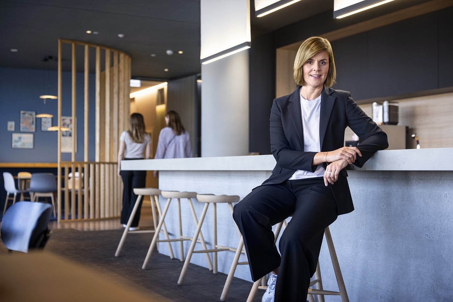 La Dra. Eva Griewel, CFO del Grupo EOS, está sentada en un espacio social en un taburete alto, vistiendo un blazer negro.